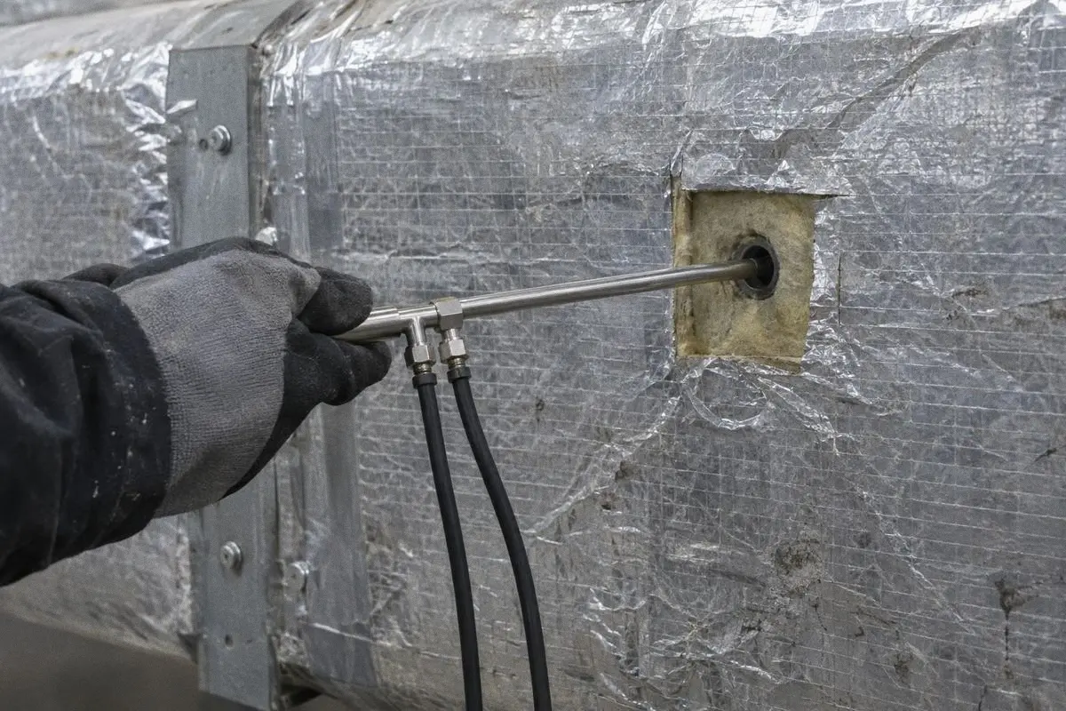 A gloved hand holds a pitot tube in a test hole on an insulated duct.